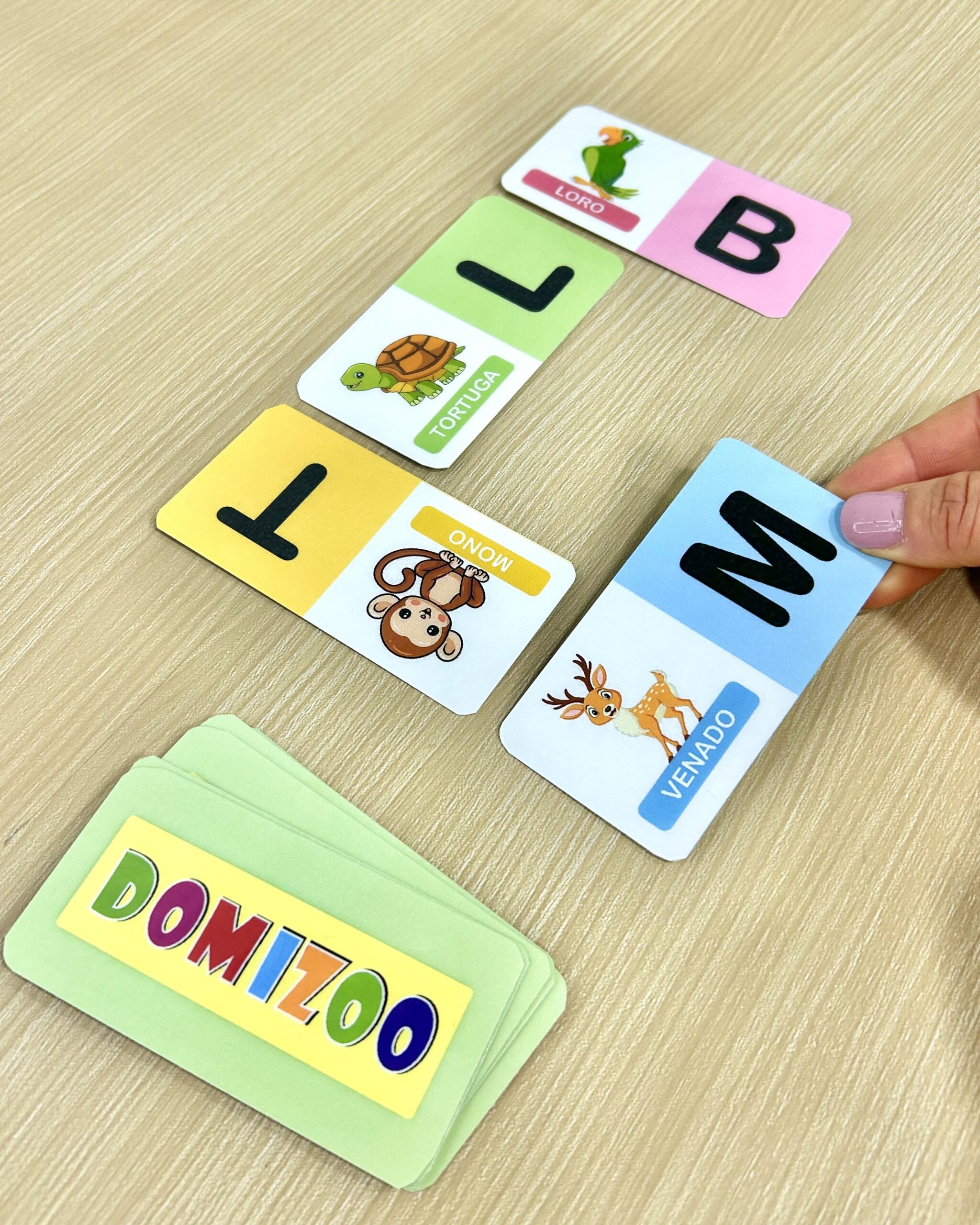 Mano colocando una tarjeta del juego Domizoo para asociar la letra M con la inicial del venado sobre mesa de madera.
