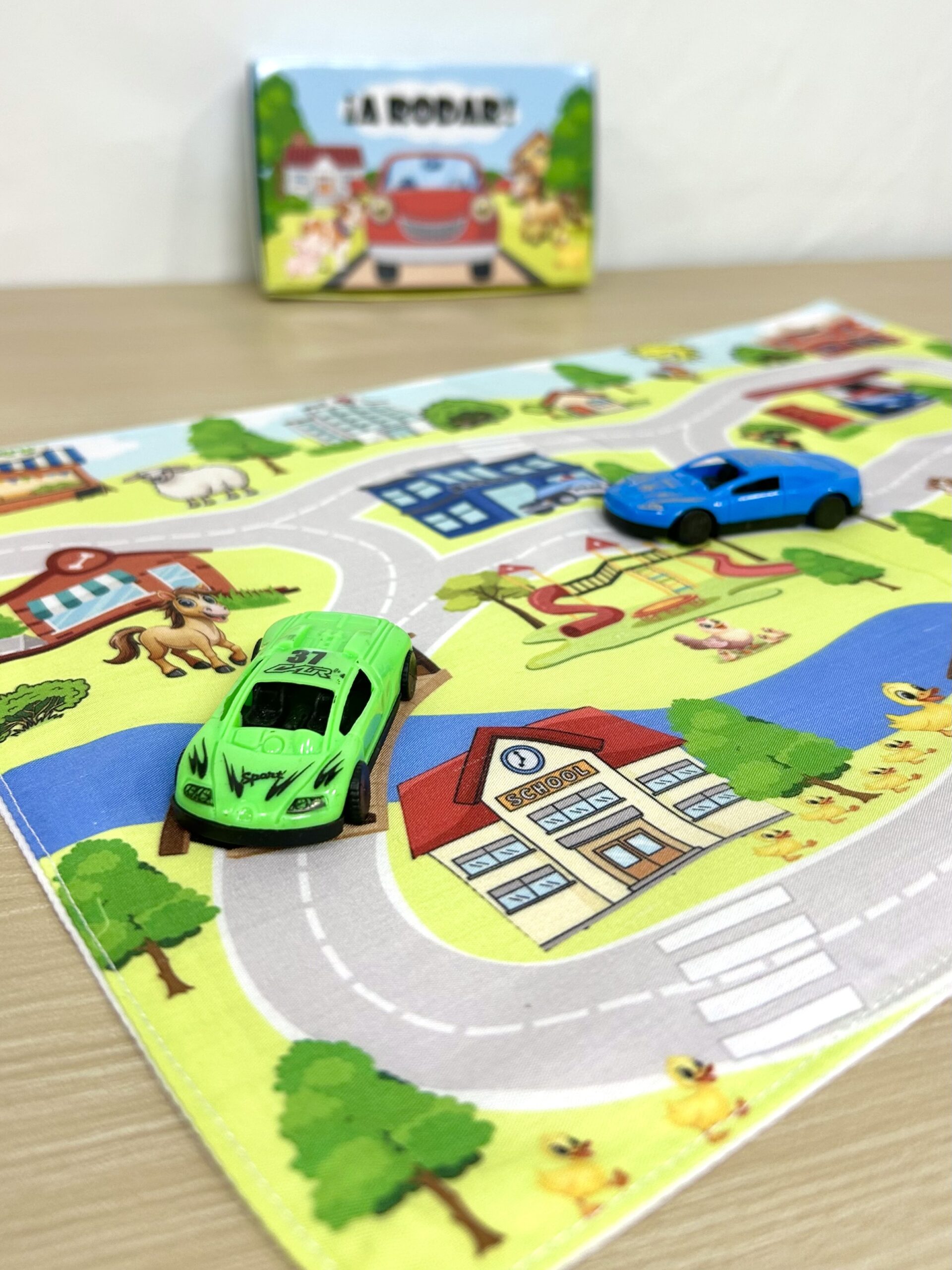 Vista de cerca de un autito de juguete verde sobre la pista de un tapiz infantil de tela impreso con ilustración de escuela y comisaría.