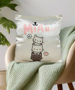Almohadón infantil satinado crema con diseño de tres gatitos kawaii colocado sobre una manta verde en un sillón moderno junto con una planta de interiores.