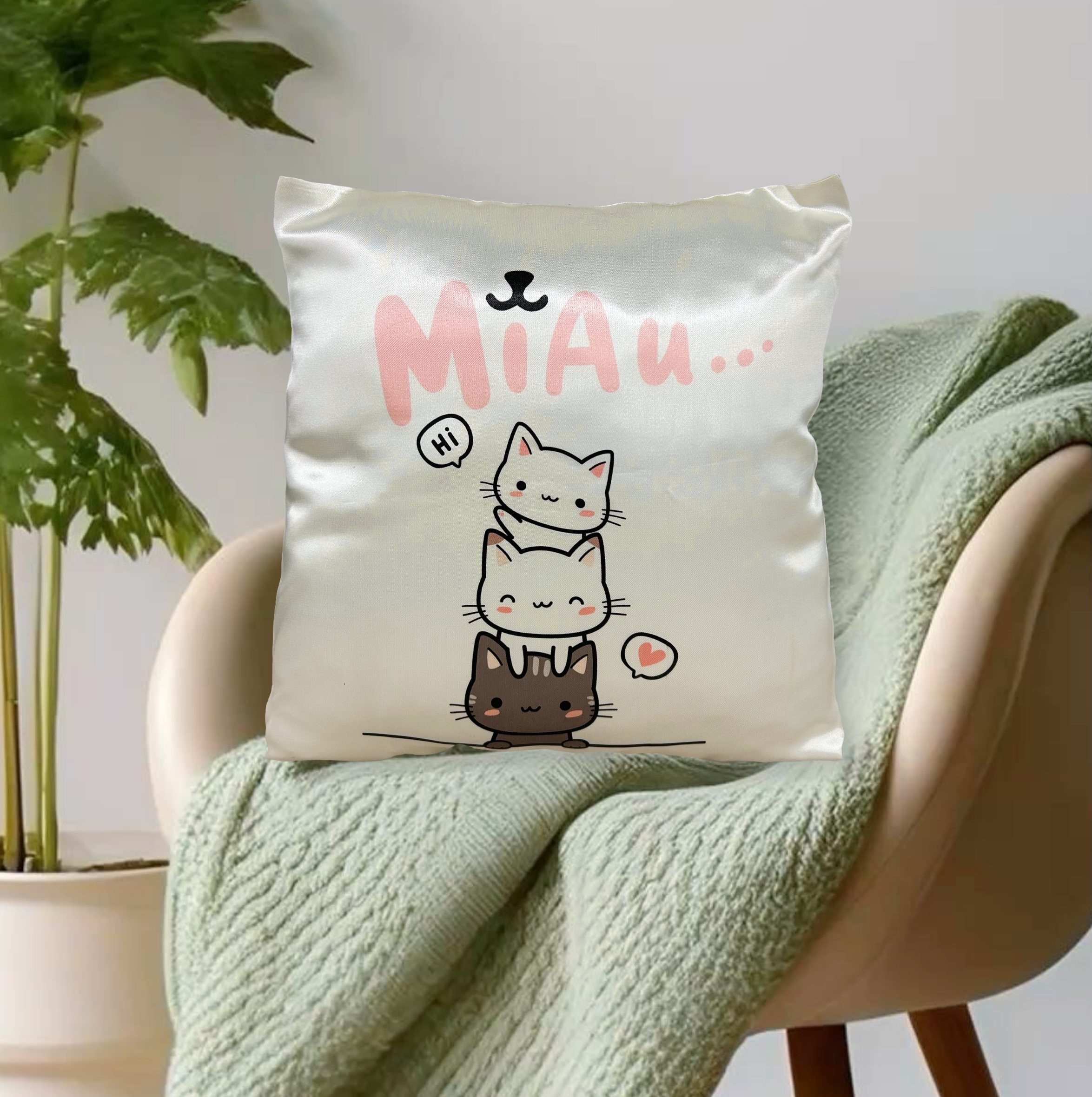 Almohadón infantil satinado crema con diseño de tres gatitos kawaii colocado sobre una manta verde en un sillón moderno junto con una planta de interiores.