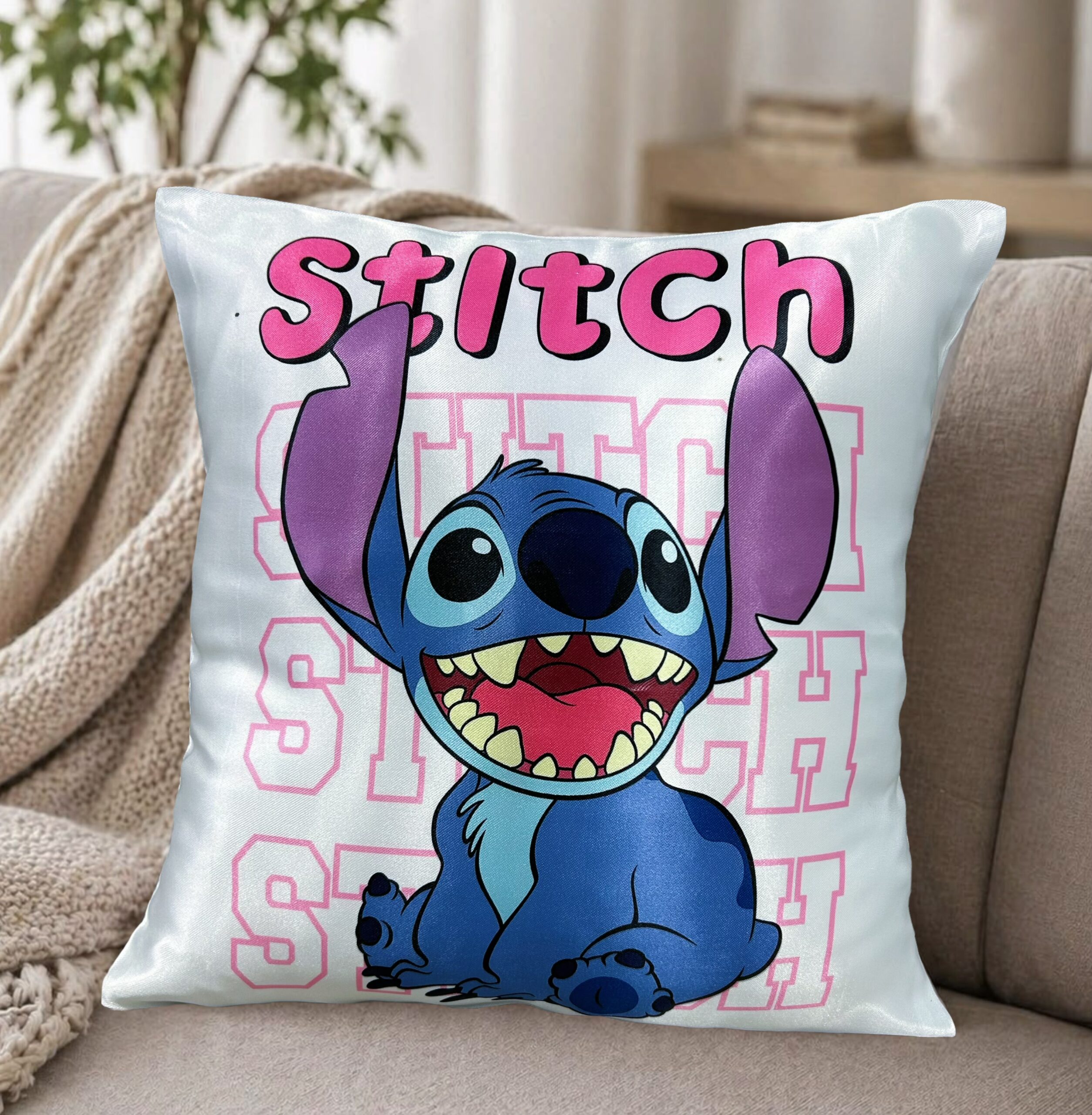 Almohadón blanco satinado con el personaje de stitch sonriendo bajo su nombre en letras rosadas, apoyado en un sillón moderno con una manta tejida de fondo.