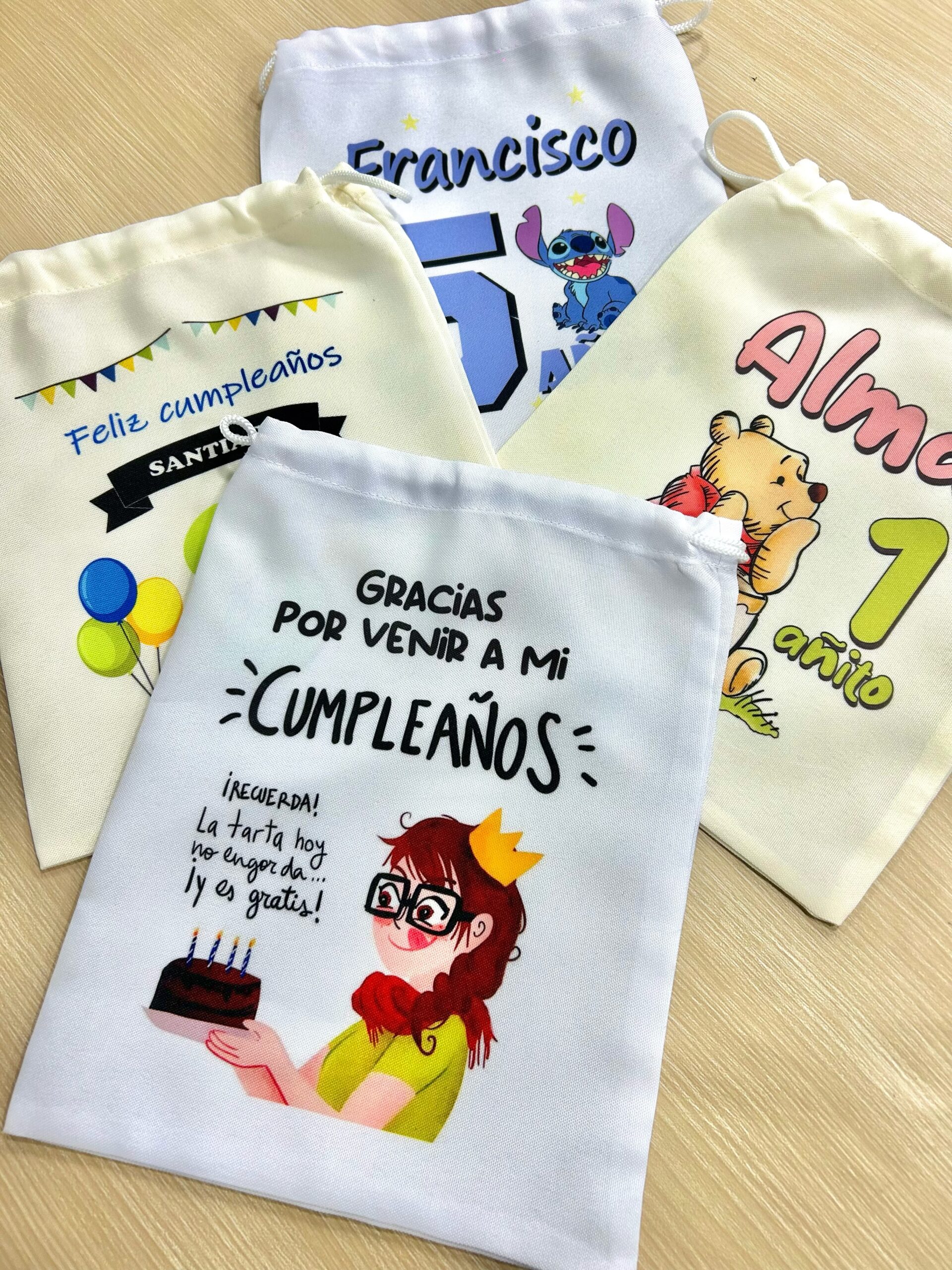 Cuatro modelos de bolsitas golosineras de tela con diferentes diseños personalizados de cumpleaños sobre mesa de madera.
